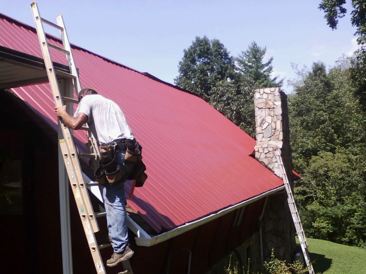 Roofer on a ladder beside a red metal roof and stone chimney — Soffit & Fascia Repair by Roofing Brothers in Newhall